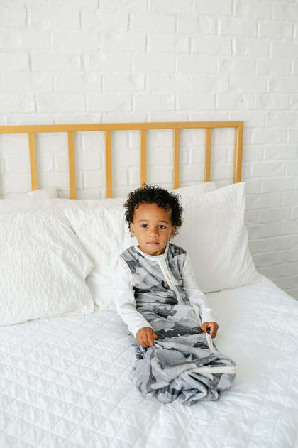 Child comfortably wearing a Camo Wearable Minky Blanket, featuring a gray camo pattern and a gold zipper, sitting on a bed.