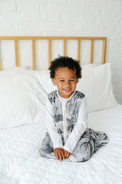 Child smiling while wearing a Camo Wearable Minky Blanket sitting on a bed with white bedding.