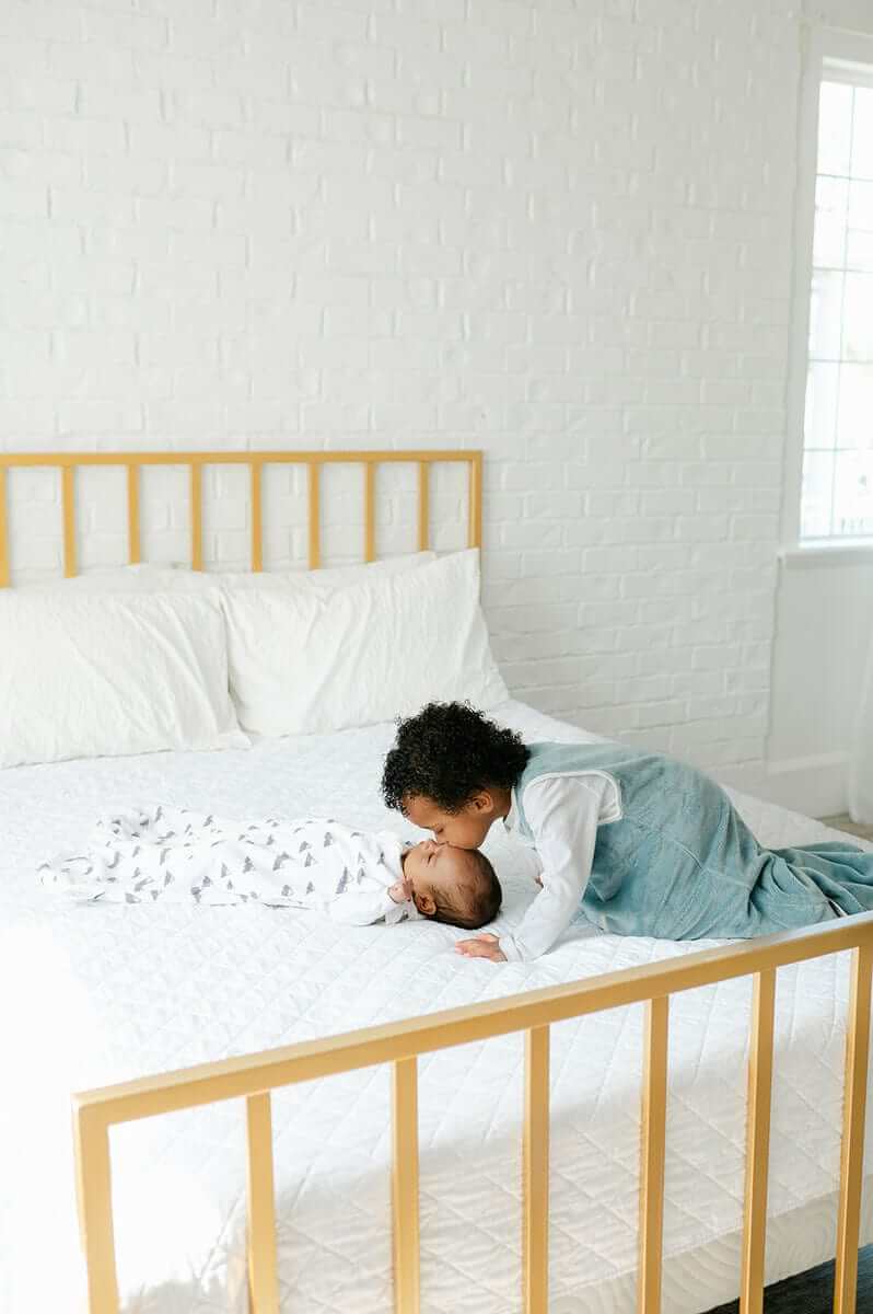 Sibling love moment with a baby on a bed, showcasing the Mountains Wearable Minky Blanket for safe infant sleep.