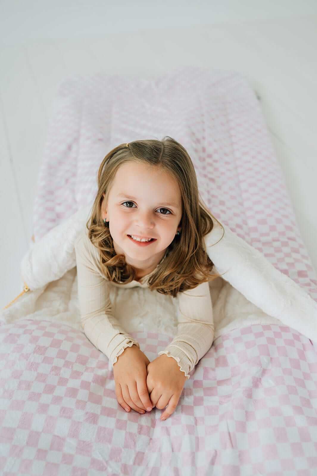Sleeping Bag in Pink Check, laid out with a smiling girl, showcasing its cozy design and plush material.