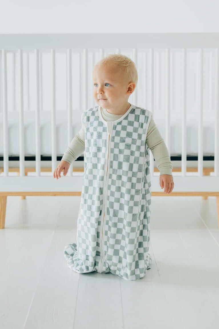 A baby wearing a Sage Check Wearable Minky Blanket standing in front of a crib, showcasing its comfy design.