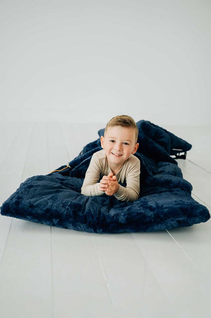 Child smiling while lying in a cozy Sleeping Bag in Navy blue, on a white floor.
