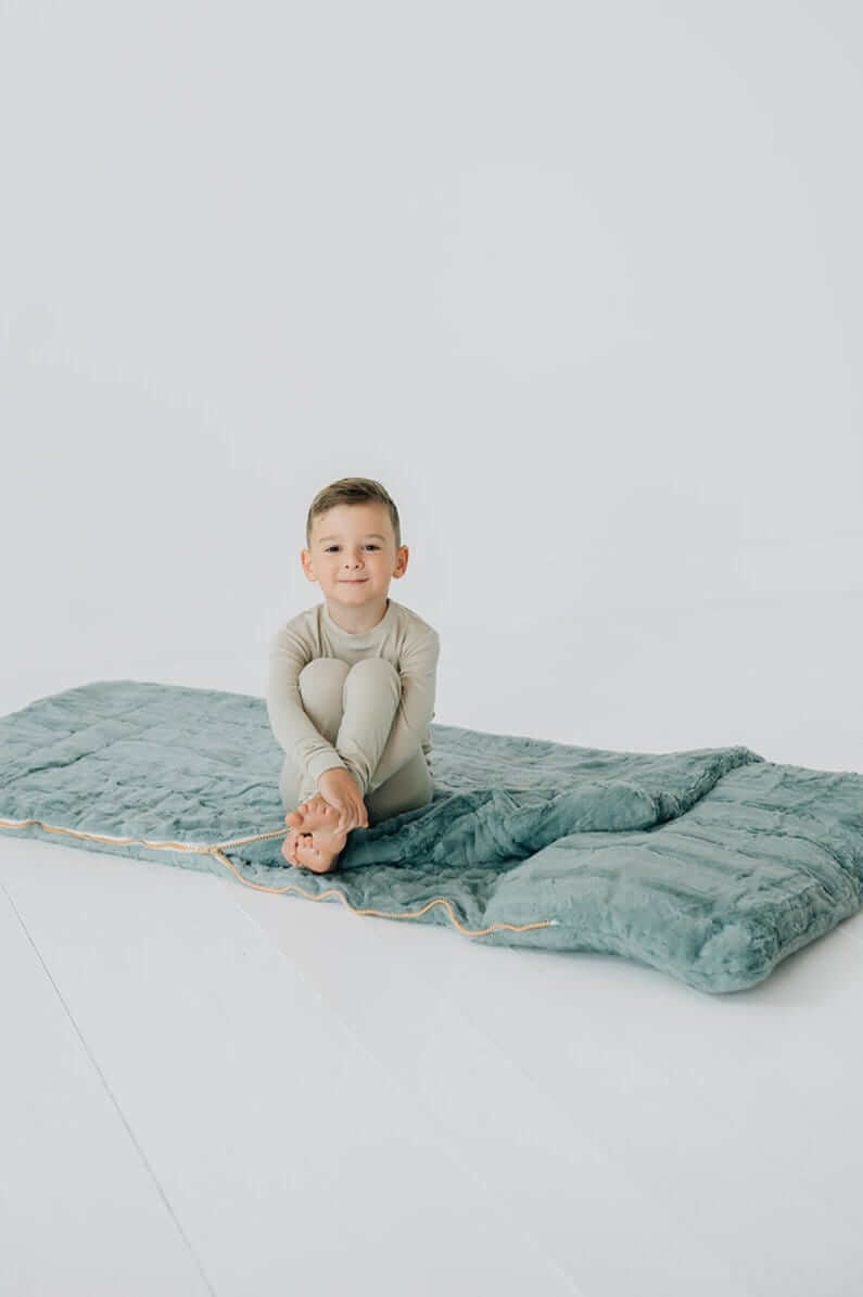 A boy sitting on a Plush Mist Sleeping Bag, showcasing its plush comfort for cozy moments.
