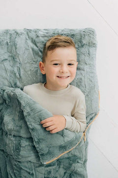 Minky Couture Plush Mist Sleeping Bag with a smiling boy cuddled inside on a cozy soft surface.