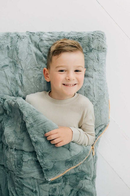 Minky Couture Plush Mist Sleeping Bag with a smiling boy cuddled inside on a cozy soft surface.