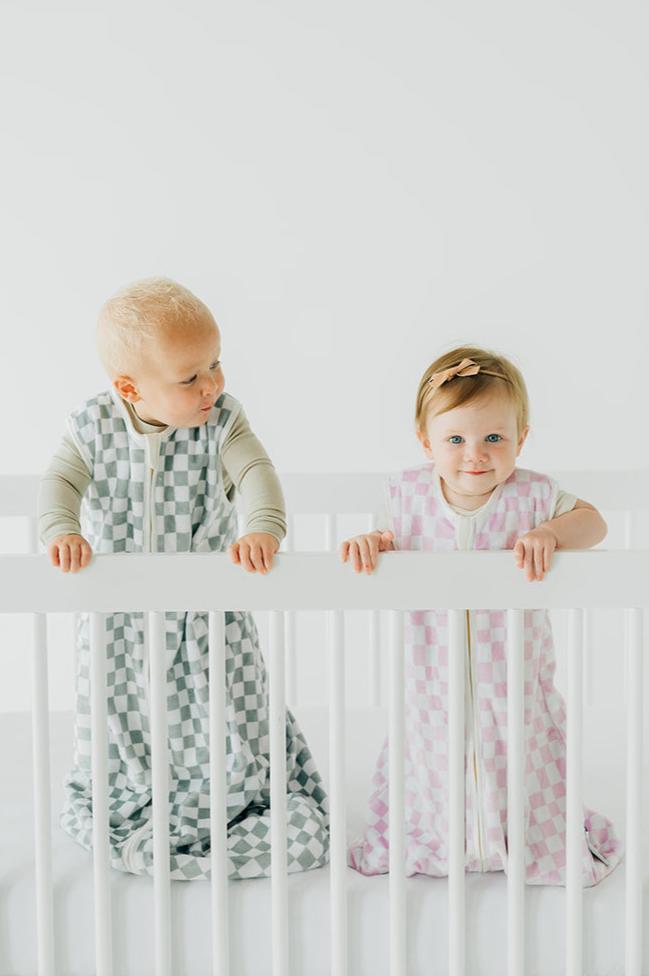 Two infants in Wearable Minky Couture blankets standing in a crib, showcasing safe sleep in stylish designs.