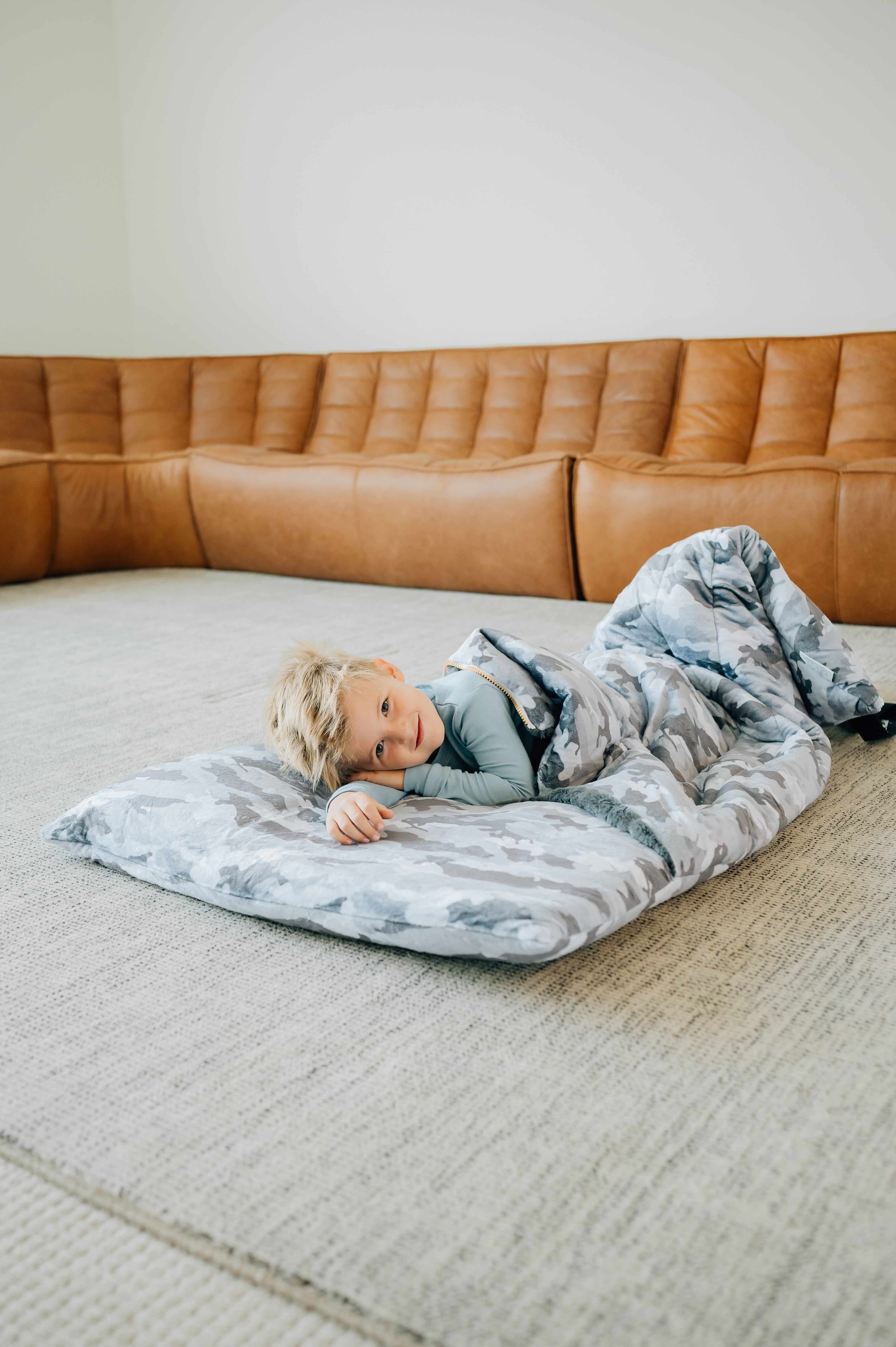 Sleeping Bag Camo featuring a child comfortably lying on a soft, plush sleeping bag in a cozy living room setting.