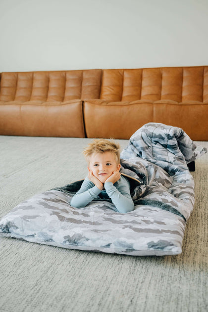 A child relaxing in a Sleeping Bag Camo on a living room floor, showcasing its plush design and comfort.