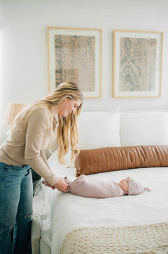 A parent lovingly wraps their baby in a Swaddle and Hat Blush set on a cozy bed.