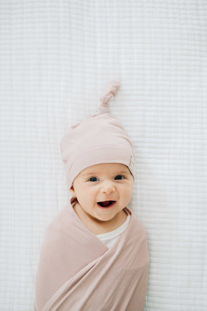 Adorable baby swaddled in soft bamboo Blush Swaddle and Hat, smiling on a cozy blanket.