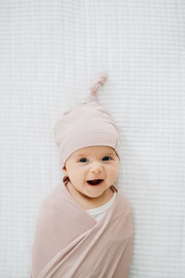 Adorable baby swaddled in soft bamboo Blush Swaddle and Hat, smiling on a cozy blanket.