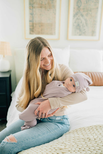 Mom cuddling baby in Swaddle and Hat Blueberry Cheesecake on a cozy bed, showcasing a stylish bonding moment.