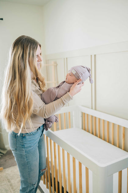 Mom cradling baby in Swaddle and Hat Blueberry Cheesecake near a crib in a cozy nursery.