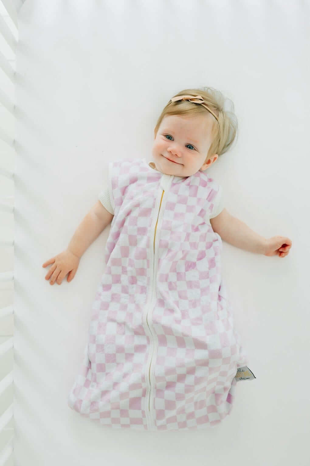 Pink Check Wearable Minky Blanket on a baby lying in a crib, showcasing its light pink check pattern and gold zipper.