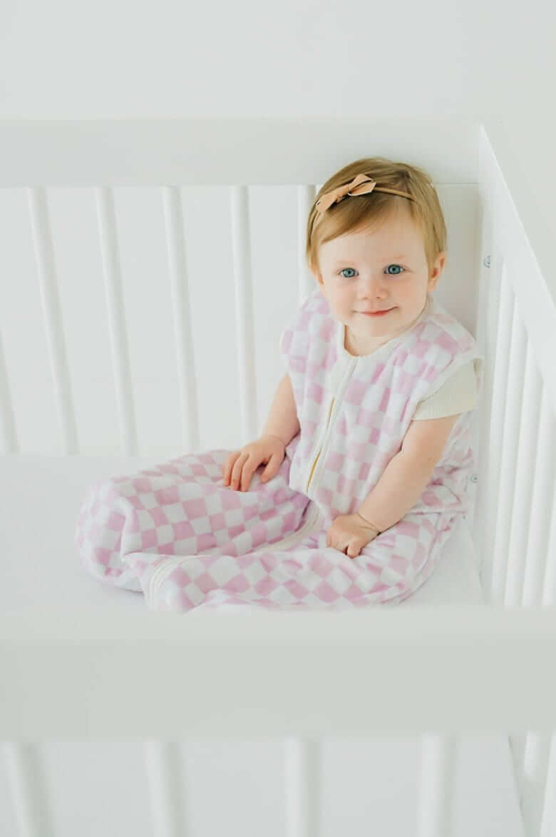 Pink Check Wearable Minky Blanket being worn by a baby sitting in a crib, showcasing its light pink check pattern.