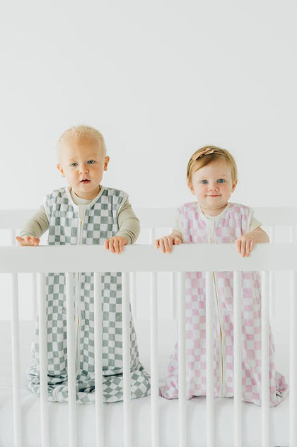 Two babies standing in a crib wearing Pink Check Wearable Minky Blanket and grey check wearable blanket.