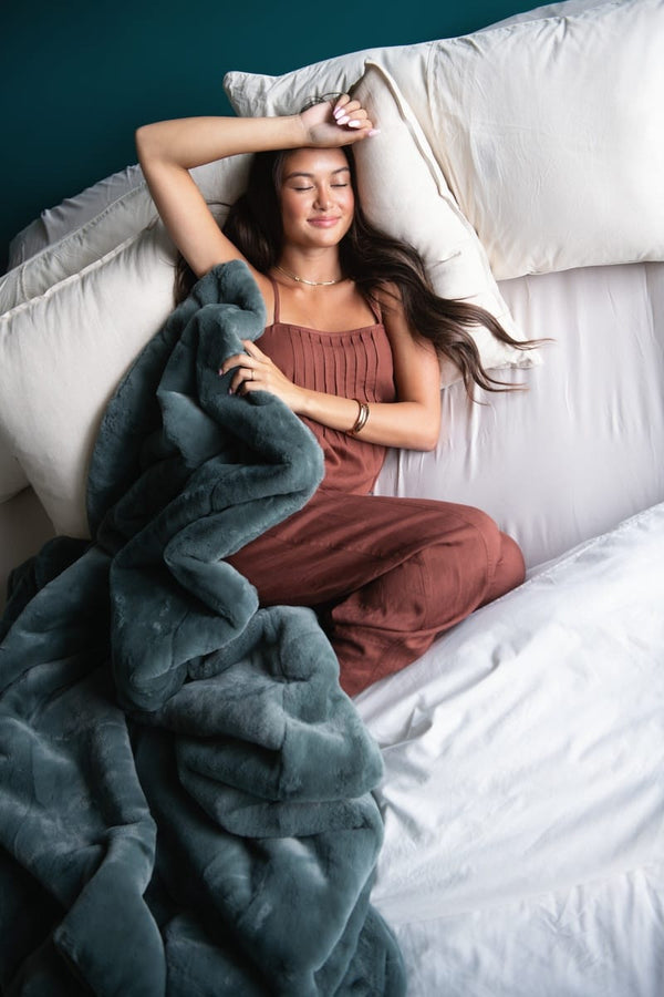 A woman relaxes on a bed with white bedding, wrapped in a plush dark teal blanket and wearing a rust-brown camisole and matching pants. She rests against white pillows against a dark teal wall.