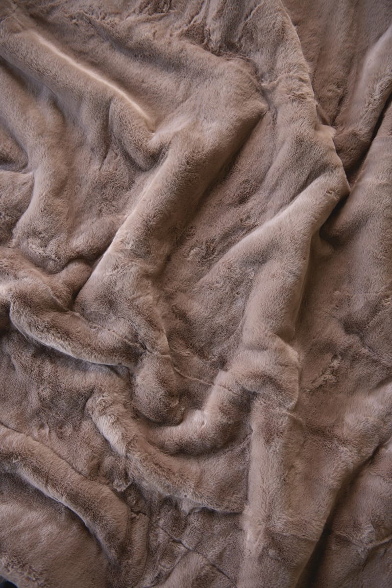 A close-up of a plush, soft faux fur or sherpa blanket in a warm taupe-brown color, shown with soft folds and ripples.
