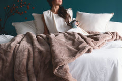 A woman sits in bed with white pillows and white bedding, wrapped in a plush taupe-brown throw blanket, holding a mug against a dark teal wall.