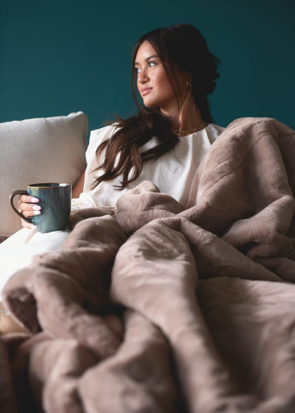A woman sits in bed under a plush taupe blanket, holding a dark teal mug against a dark teal wall background.