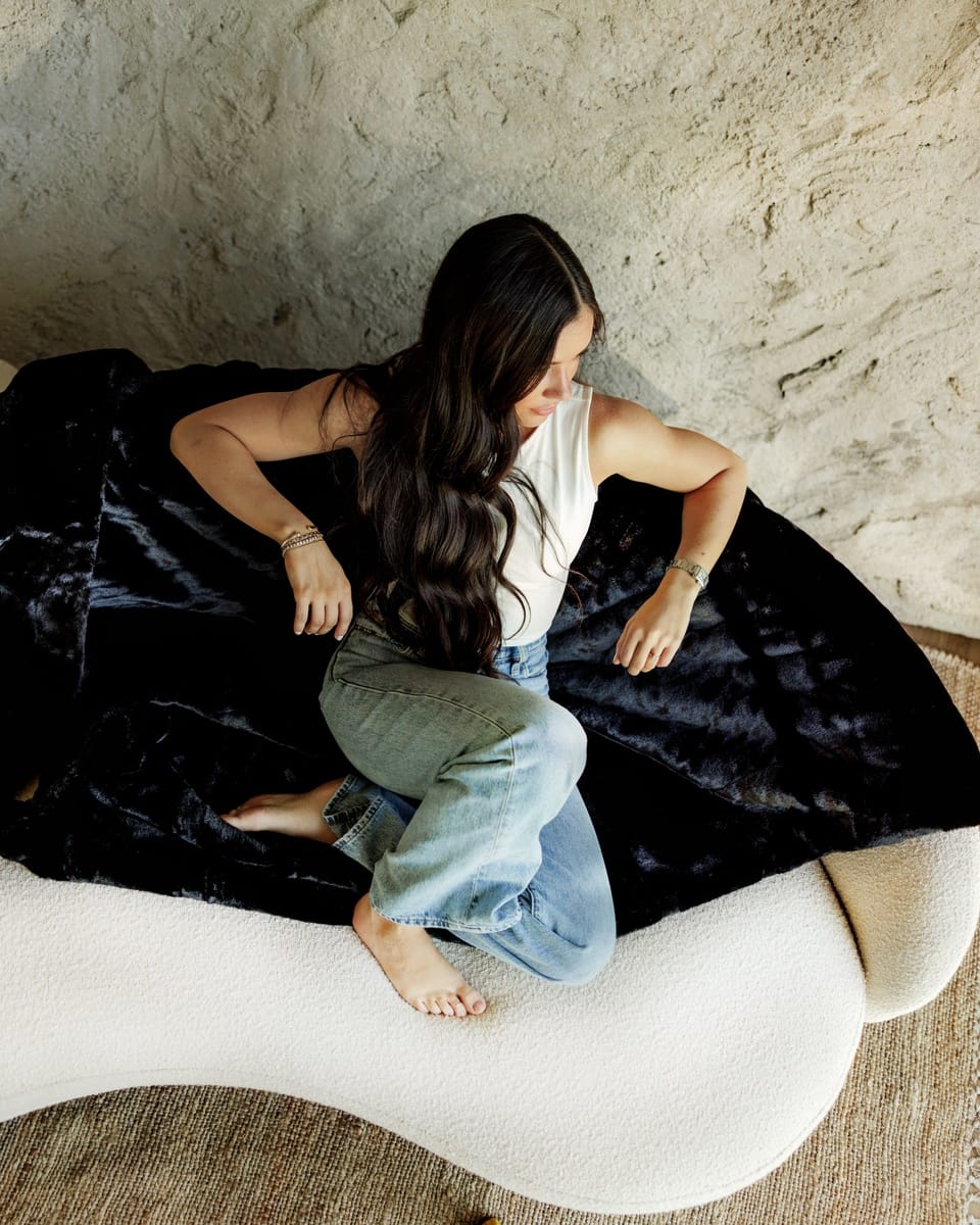 A woman sits barefoot on a cream-colored curved bench, wearing a white tank top and light blue jeans, with a Lush Black Onyx Blanket draped behind her against a textured beige wall.