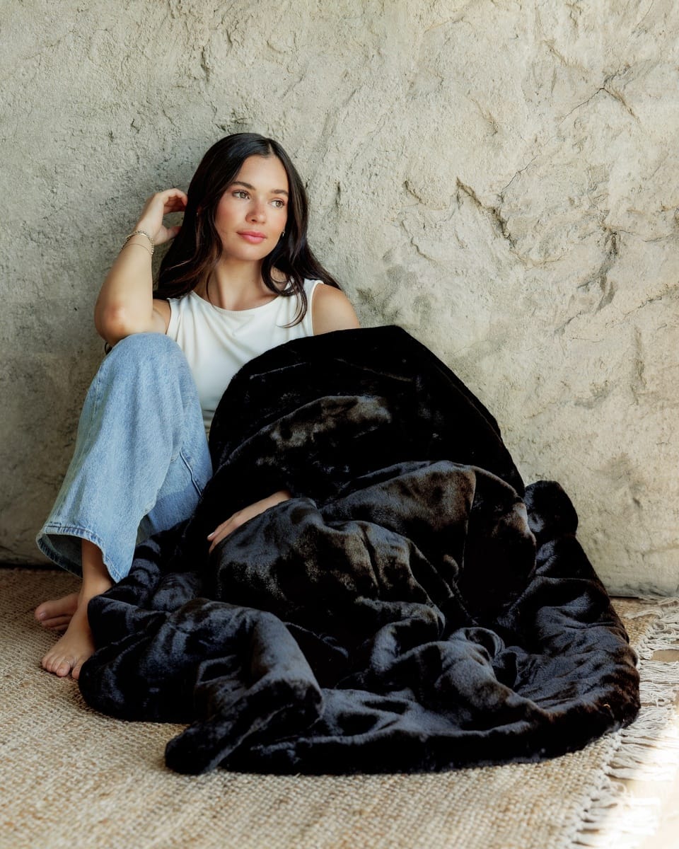 A woman sits barefoot against a textured plaster wall, wrapped in a Lush Black Onyx Blanket. She is wearing a white tank top and light blue jeans.