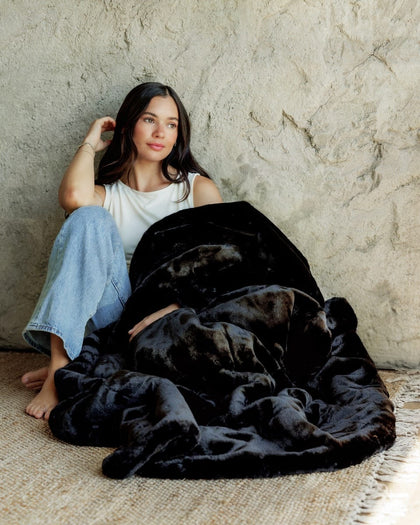 A woman sits barefoot against a textured plaster wall, wrapped in a Lush Black Onyx Blanket. She is wearing a white tank top and light blue jeans.