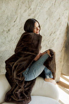 A woman sits barefoot on a white upholstered chair wrapped in a plush, Lush Espresso Blanket against a textured light beige wall.