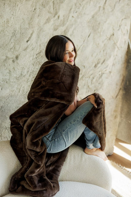 A woman sits barefoot on a white upholstered chair wrapped in a plush, Lush Espresso Blanket against a textured light beige wall.