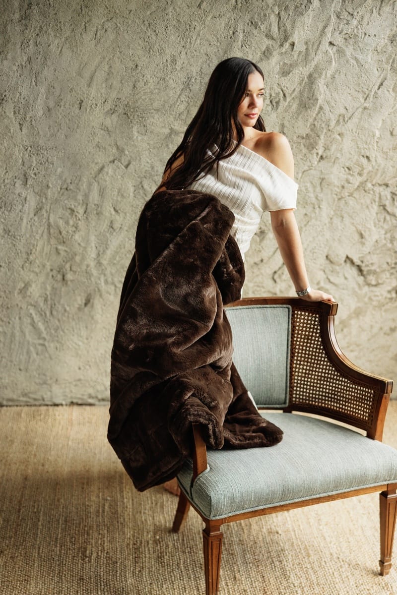 A woman with long dark hair sits on a light blue upholstered wooden chair, wrapped in a plush Lush Espresso Blanket. She is wearing a white off-the-shoulder top.