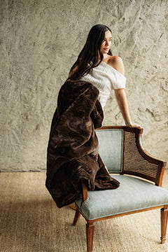A woman with long dark hair sits on a light blue upholstered wooden chair, wrapped in a plush Lush Espresso Blanket. She is wearing a white off-the-shoulder top.