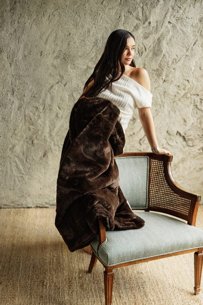 A woman with long dark hair sits on a light blue upholstered wooden chair, wrapped in a plush Lush Espresso Blanket. She is wearing a white off-the-shoulder top.