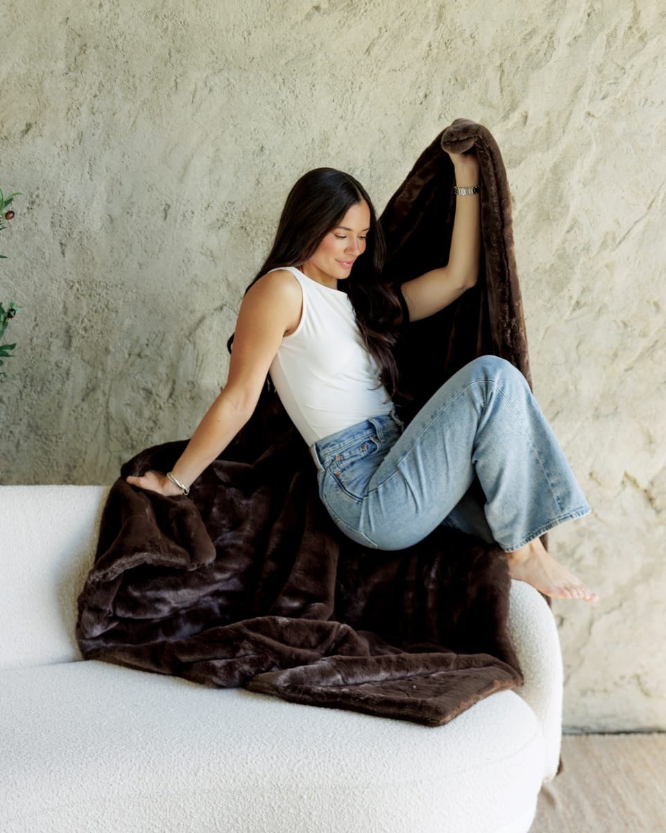 A woman sits on a white sofa draped in a plush dark brown Lush Espresso Blanket, holding up the opposite corner of the blanket against a textured beige wall.