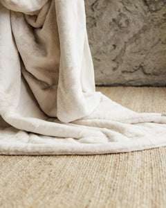 A soft, plush Lush Macadamia Nut Blanket draped over a light beige woven rug in front of a textured gray wall.