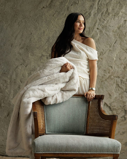 A woman with long dark hair sits on a wooden armchair draped in a Lush Macadamia Nut Blanket, wearing a white off-the-shoulder dress against a textured gray wall.