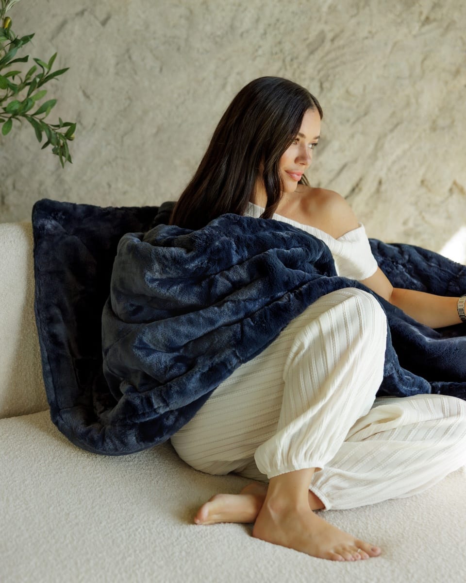 A woman sits barefoot on a textured white sofa, wrapped in a plush navy blue minky couture blanket. She is wearing a white off-the-shoulder top and white lounge pants, with a neutral textured wall and a small green plant in the background.