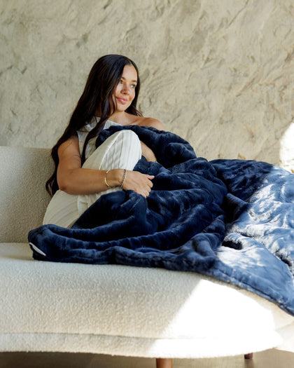A woman sits on a cream boucle sofa wrapped in a plush navy blue Minky Couture  blanket, with sunlight streaming across the scene.