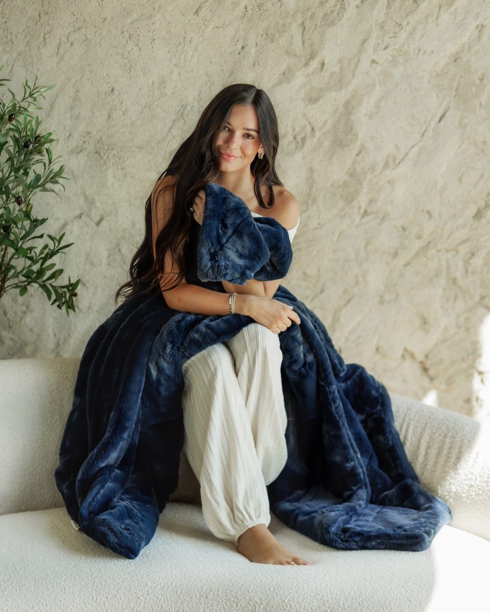 A woman sits barefoot on a white textured sofa, wrapped in and holding a plush navy blue Minky Couture Lush Night Sky blanket against a light beige stucco wall.