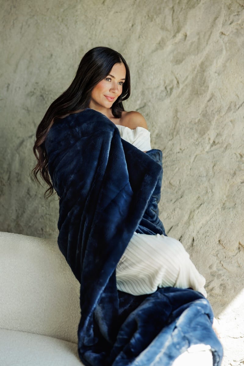A woman sits on a light-colored sofa wrapped in a plush navy blue lush minky couture blanket, with a textured beige wall behind her.
Details
