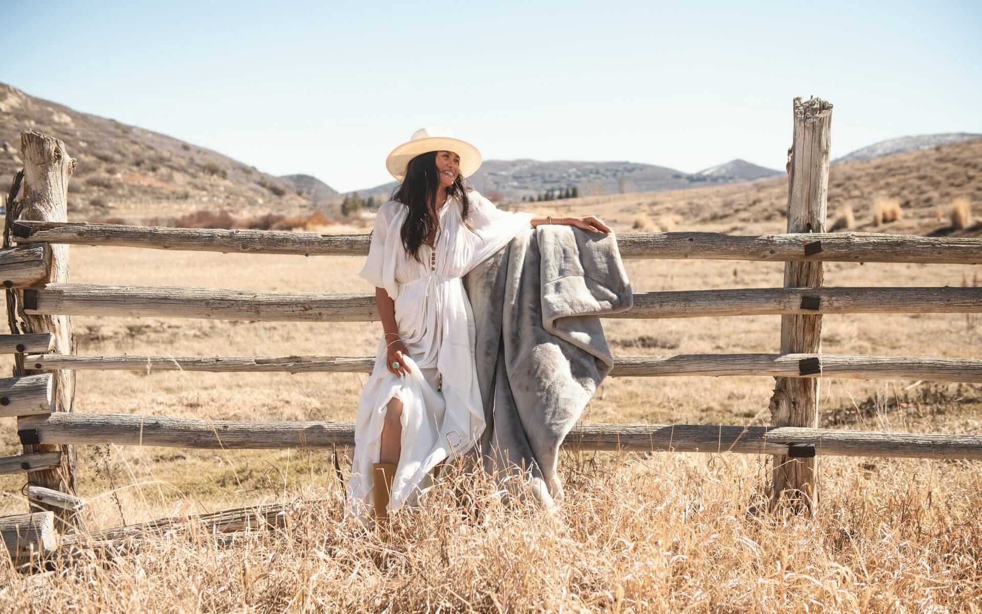 A woman in a white dress and hat holding the Lush Putty Blanket while standing by a wooden fence in a rustic landscape.