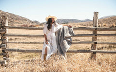 A woman in a white dress and hat holding the Lush Putty Blanket while standing by a wooden fence in a rustic landscape.