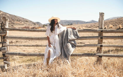 A woman in a white dress and hat holding the Lush Putty Blanket while standing by a wooden fence in a rustic landscape.