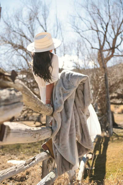 A woman draped in a Lush Putty Blanket, wearing a white dress and hat, enjoying the outdoors.
