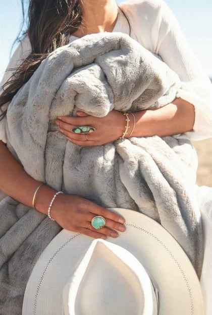 A woman wrapped in a Lush Putty Blanket holds it close, showcasing its soft gray fabric and elegant design.