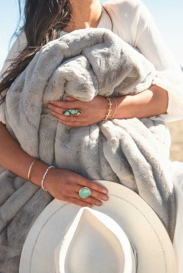 A woman wrapped in a Lush Putty Blanket holds it close, showcasing its soft gray fabric and elegant design.