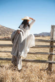Woman in a white dress holding Lush Putty Blanket while sitting on a wooden fence in a scenic outdoor setting.