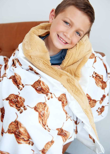 A smiling boy cuddled in a Highland Cow Blanket featuring highland cows, with a plush backing and cream ruffle.