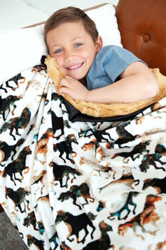 A boy smiling while wrapped in a Stallion Blanket featuring watercolor horses on a cream background.