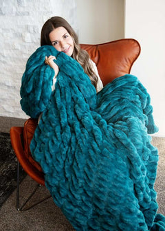 Woman wrapped in the cozy Hugs Deep Lake Blanket, enjoying plush comfort while seated on a stylish chair.