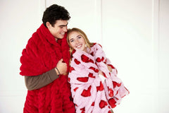 Couple snuggled in Hugs Heart Red Blanket, featuring vibrant red hearts on a soft pink background.